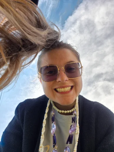 Ana de óculos escuros, fotografada de baixo com o céu azul e nuvens ao fundo